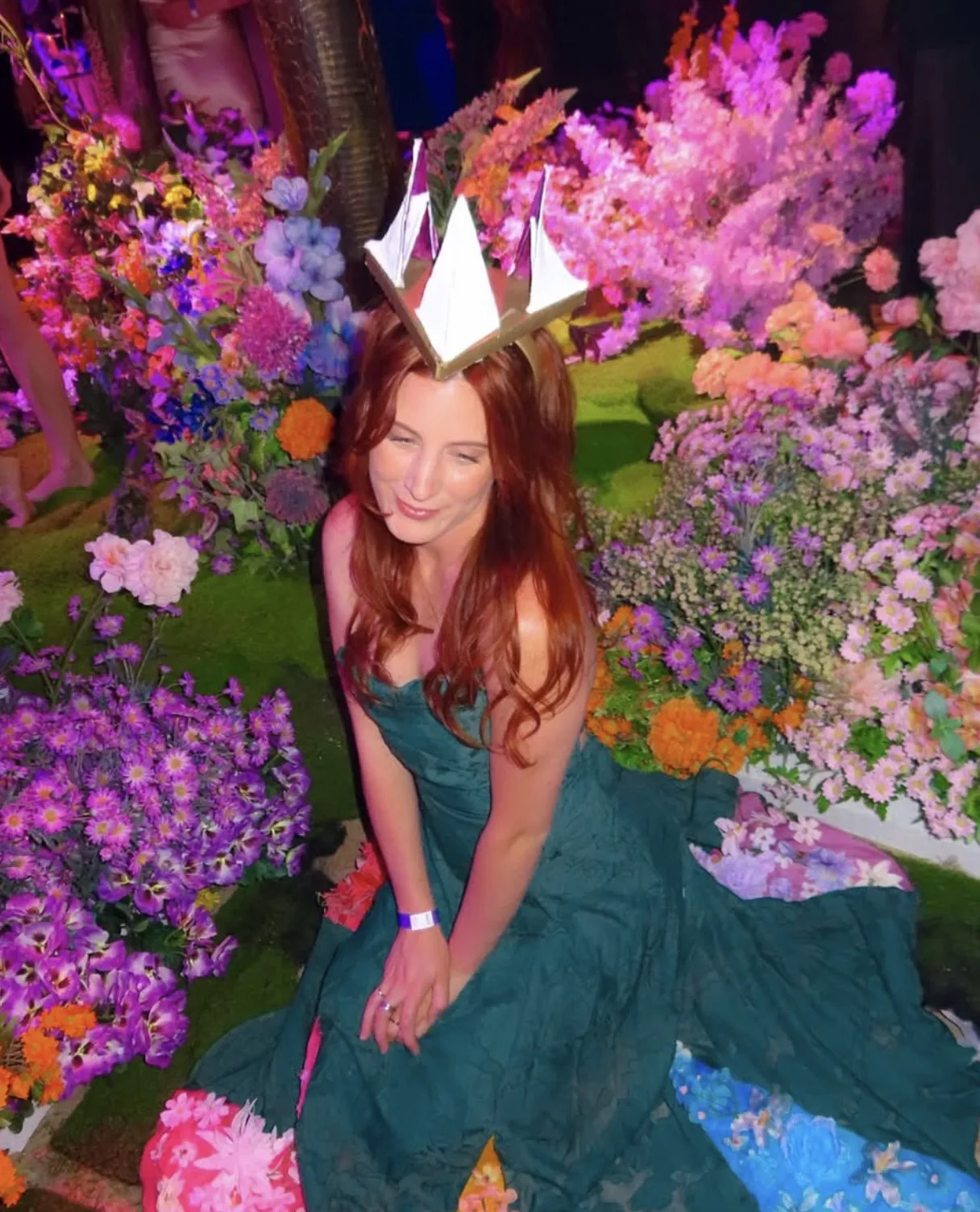 Arabella Rosier in a green lace gown and a folded paper crown, sitting among banks of pink, purple and orange flowers on a moss floor