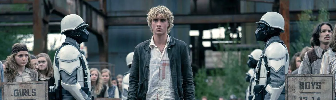 Joseph Zada as Haymitch Abernathy walks past Peacekeepers and the 12 to 14 age pens at the District 12 reaping