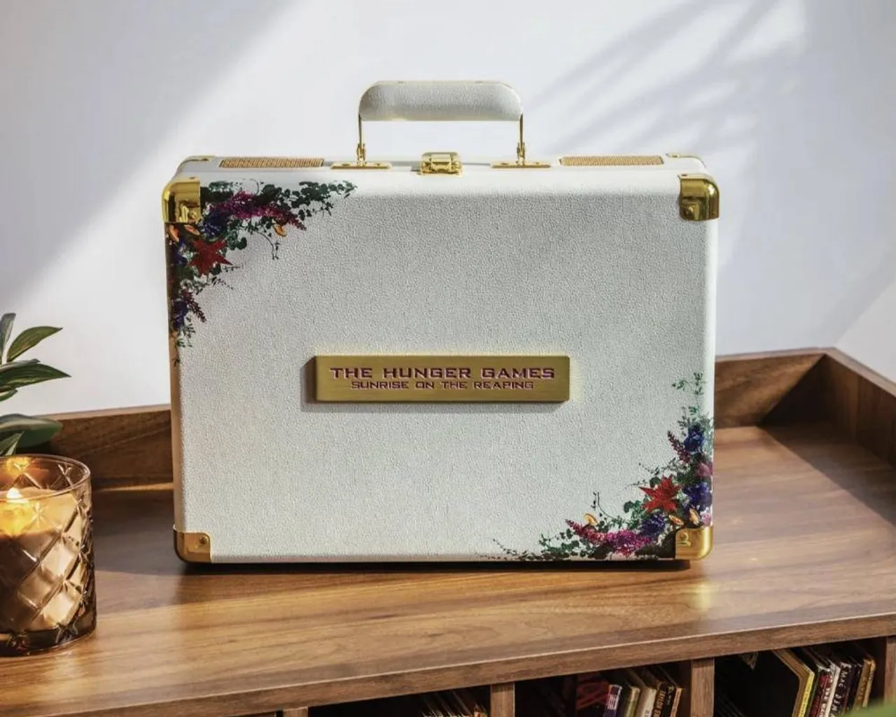 The limited edition Hunger Games Crosley Cruiser closed on a timber record cabinet: a pebbled white case with gold corners, wildflowers on two corners and a gold plate reading The Hunger Games, Sunrise on the Reaping