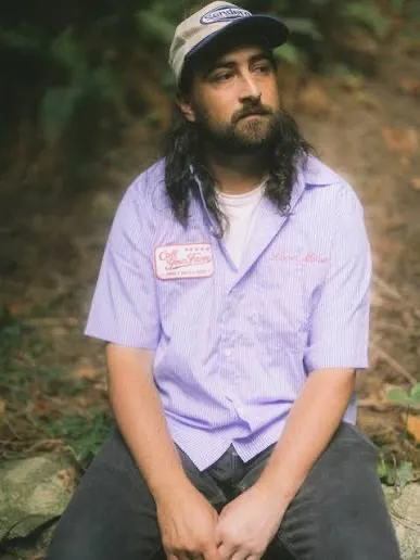 Noah Kahan in a lilac short-sleeved work shirt and cap, seated on a rock outdoors