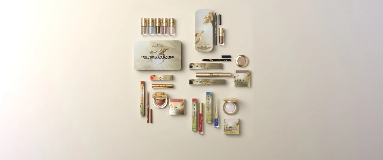 The full Stila × The Hunger Games: Sunrise on the Reaping collection laid out overhead: the gold Sunrise Eyes tin, the All Fire duo, five Liqua-Play liquid eyeshadows, liners, mascara, highlighters and lipstick, all in gold packaging with the snake-and-songbird motif