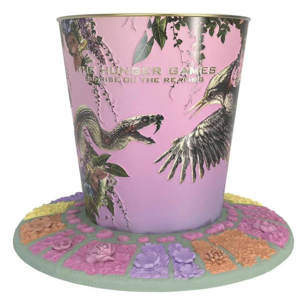 Village Cinemas' Sunrise on the Reaping popcorn tub: pink, with a snake and a mockingjay among trailing vines, sitting on a round sage-green base covered in pink, orange, yellow and lilac flowers.